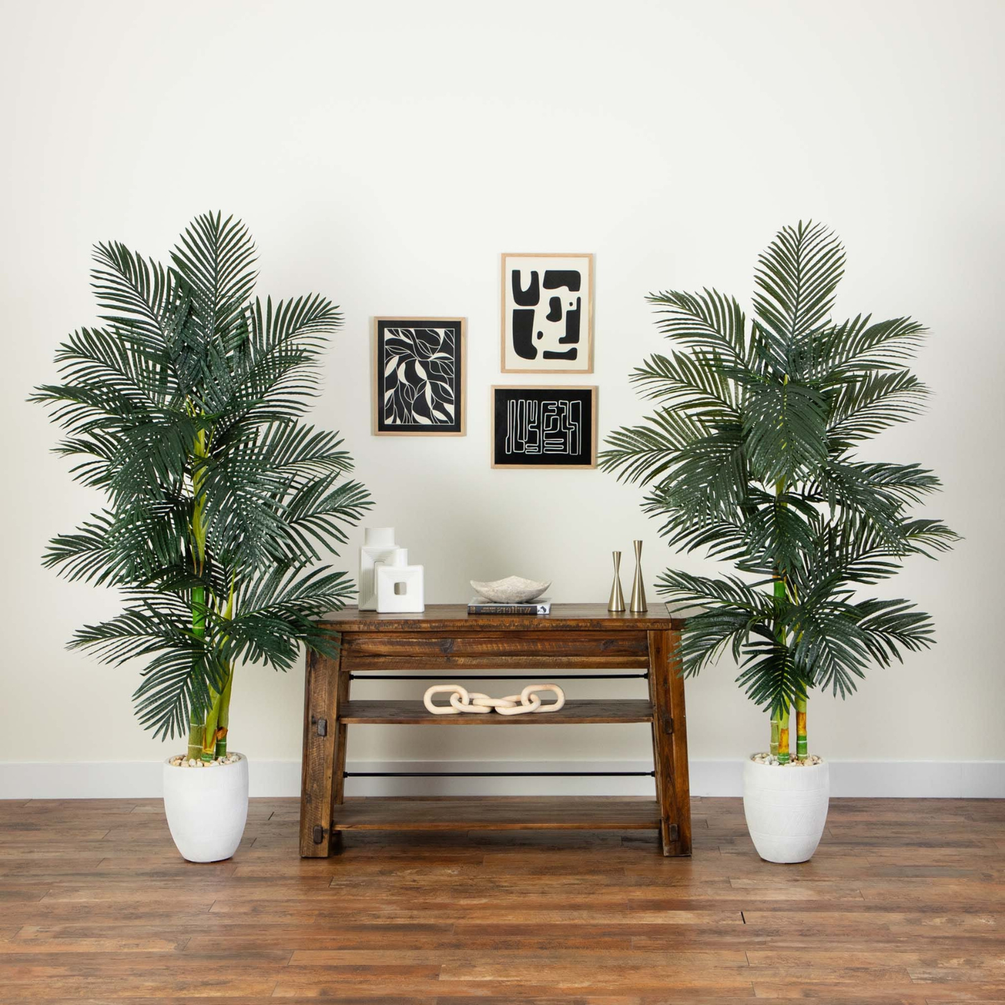 Two tall artificial palm trees in white planters flanking a dark wood console table against a white wall with framed abstract art.
