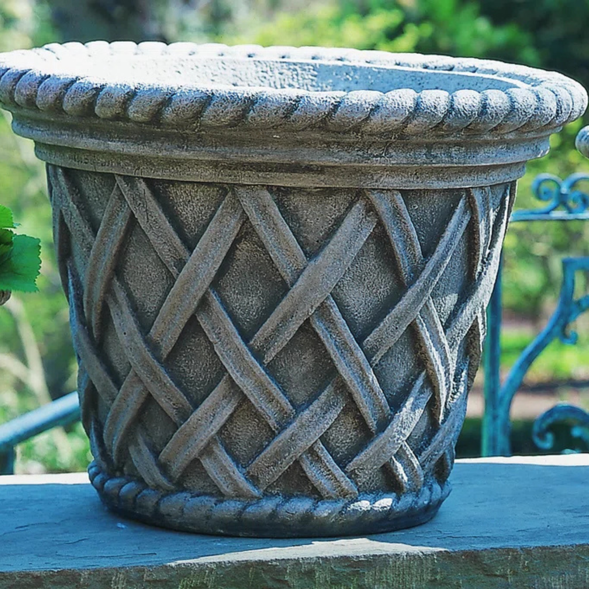 A round stone-look planter pot with a textured woven basket pattern and a rope-trimmed rim, shown outdoors on a stone ledge with blurred greenery and a blue metal railing in the background.