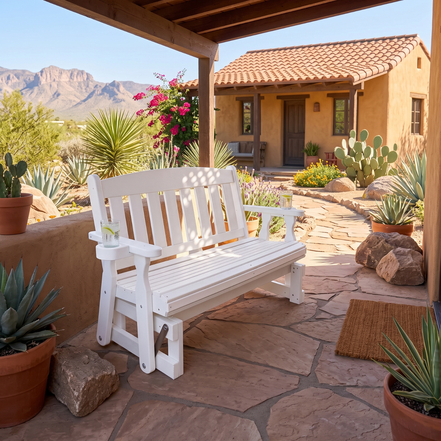A white wooden outdoor bench with a slatted back and seat, placed on a stone patio under a covered porch with desert landscaping, cacti, and a stucco house in the background.