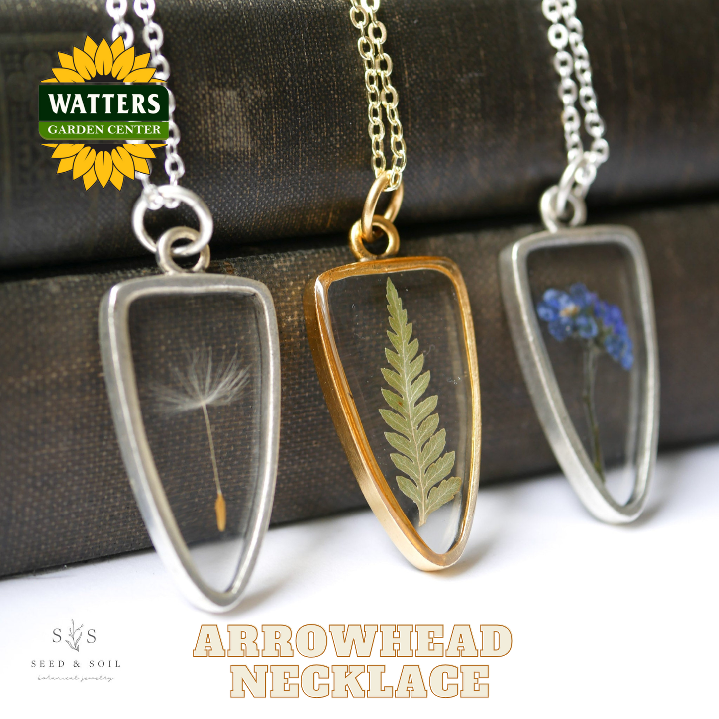 Three arrowhead necklaces with pressed flowers and plants on a brown surface, featuring Watters Garden Center logo.