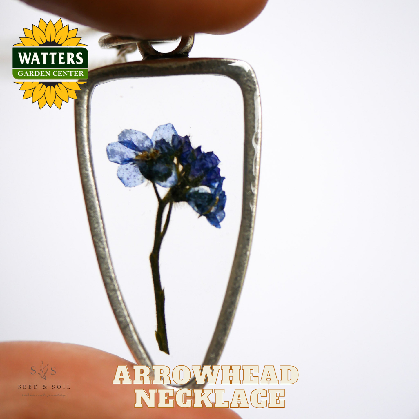 Silver arrowhead necklace with a blue flower pendant held by a hand, Watters Garden Center logo visible.
