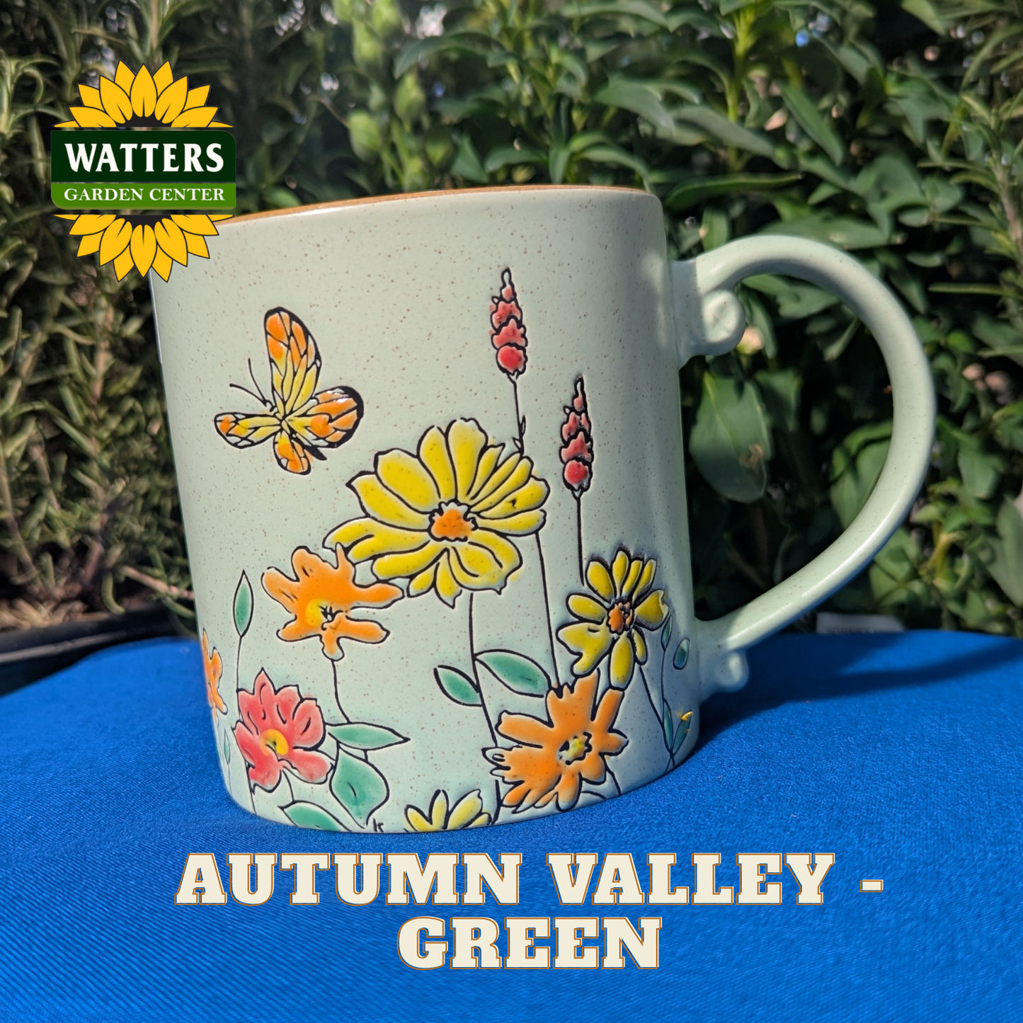 Butterfly and floral pattern on a green mug with blue background behind the Autumn Valley - Green mug with Watters logo.