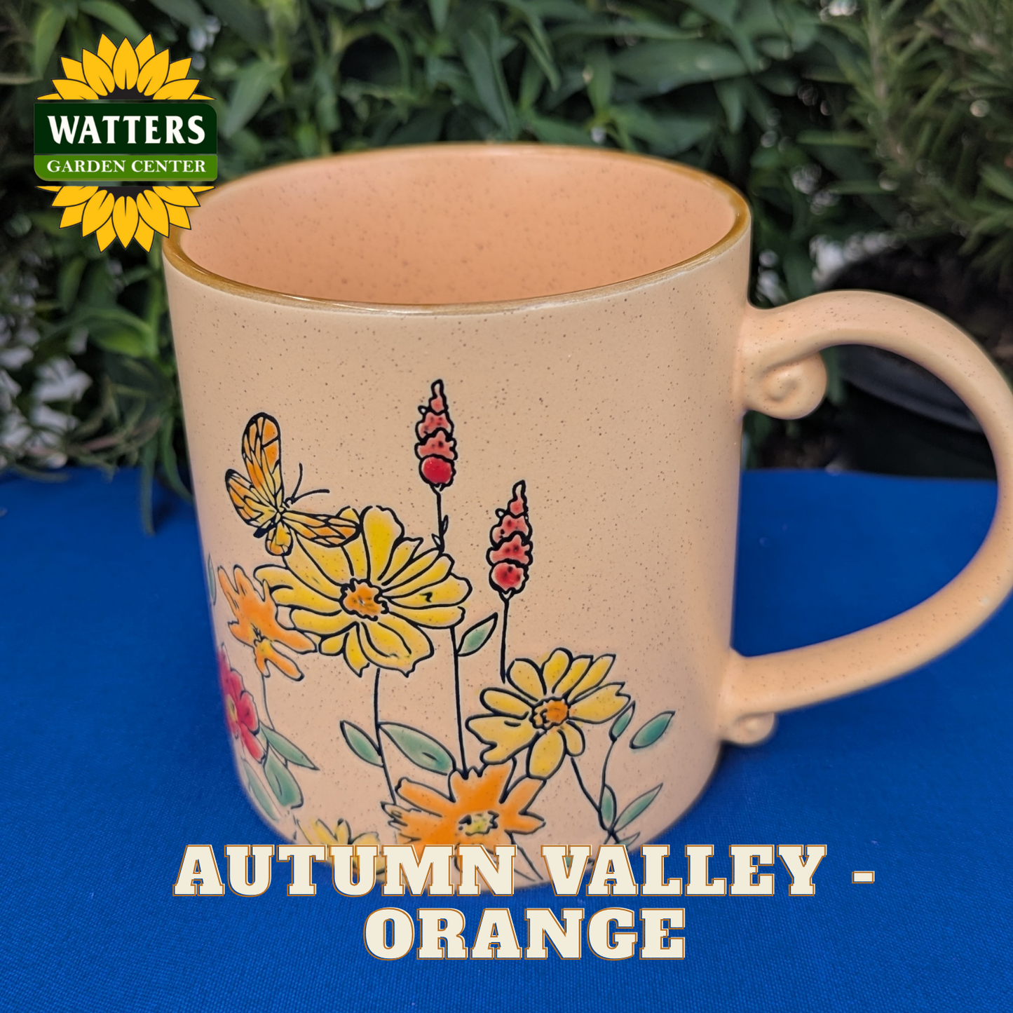 Beige mug with floral design on a blue surface, featuring Watters Garden Center branding.