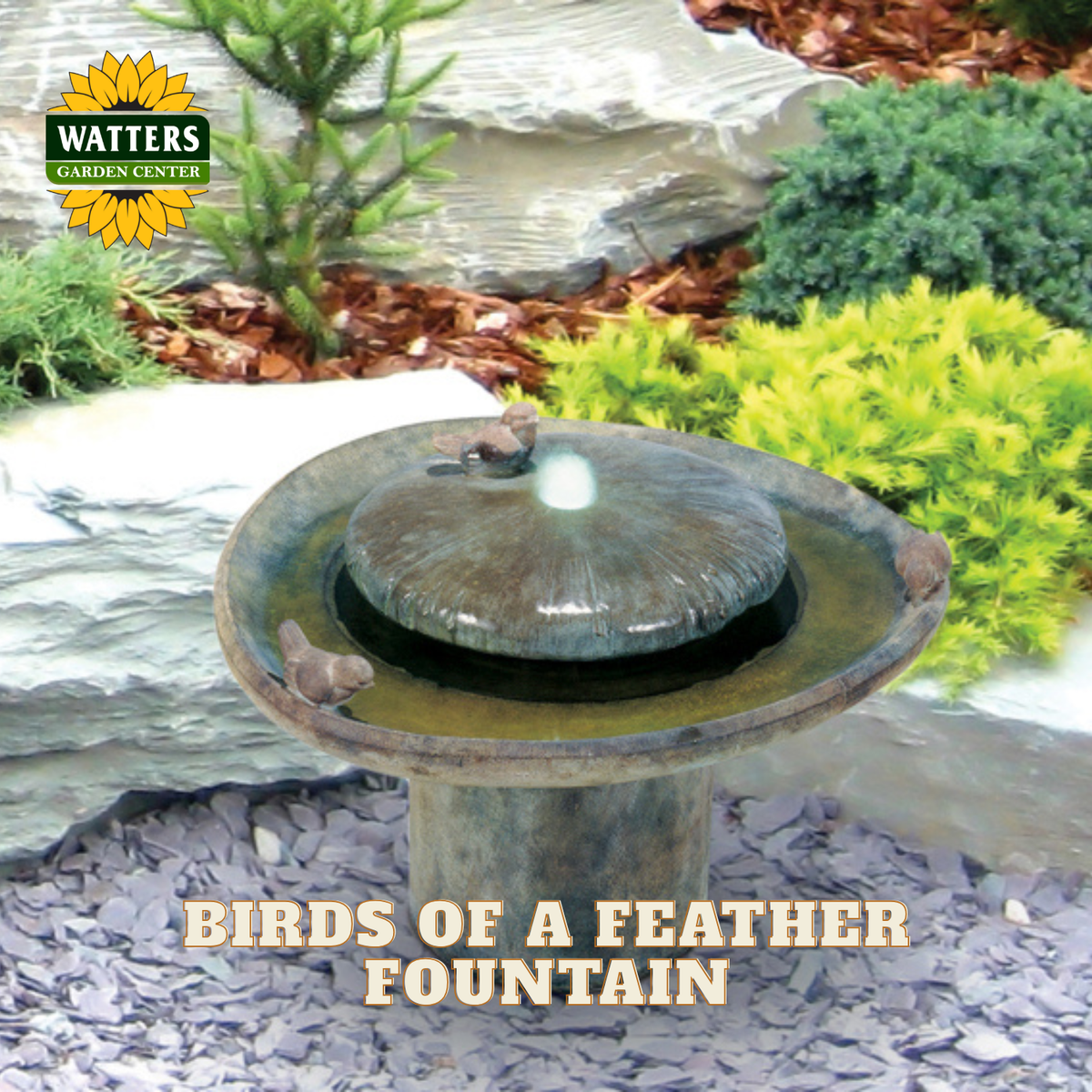 Watters Garden Center Birds of a Feather Fountain shown in a landscaped garden setting with rocks, shrubs, and mulch. The fountain has a stone-textured base and lid with small bird figurines perched on top, and water is visibly flowing from the center.