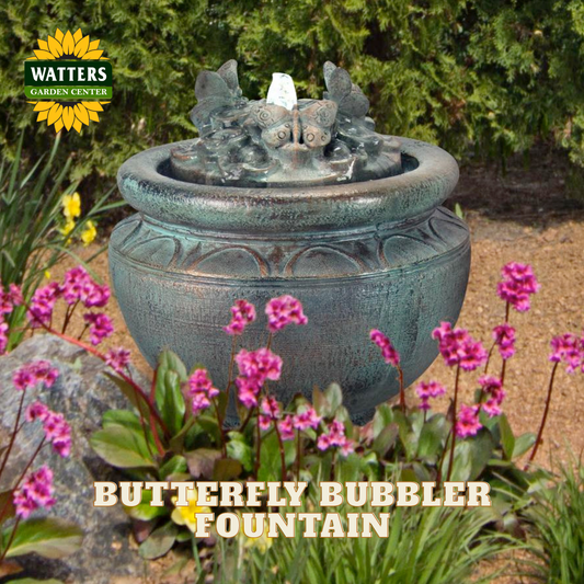 A Watters Garden Center Butterfly Bubbler Fountain in a garden setting, featuring a textured teal-green stone-like pot with a sculpted butterfly spout and flowing water, surrounded by pink flowers and green foliage.