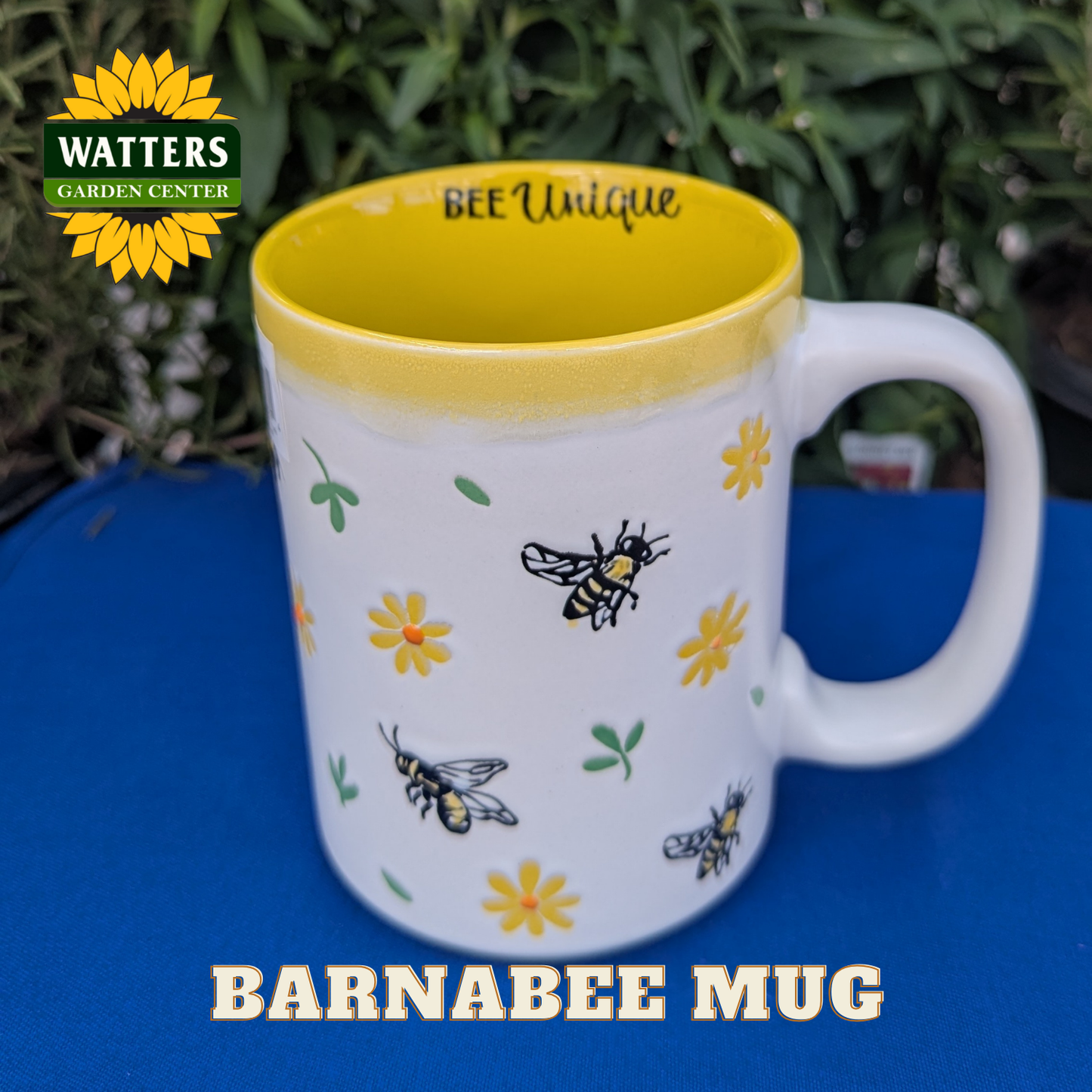 White mug with bee and flower design on a yellow rim, placed on a blue surface with Watters Garden Center branding.