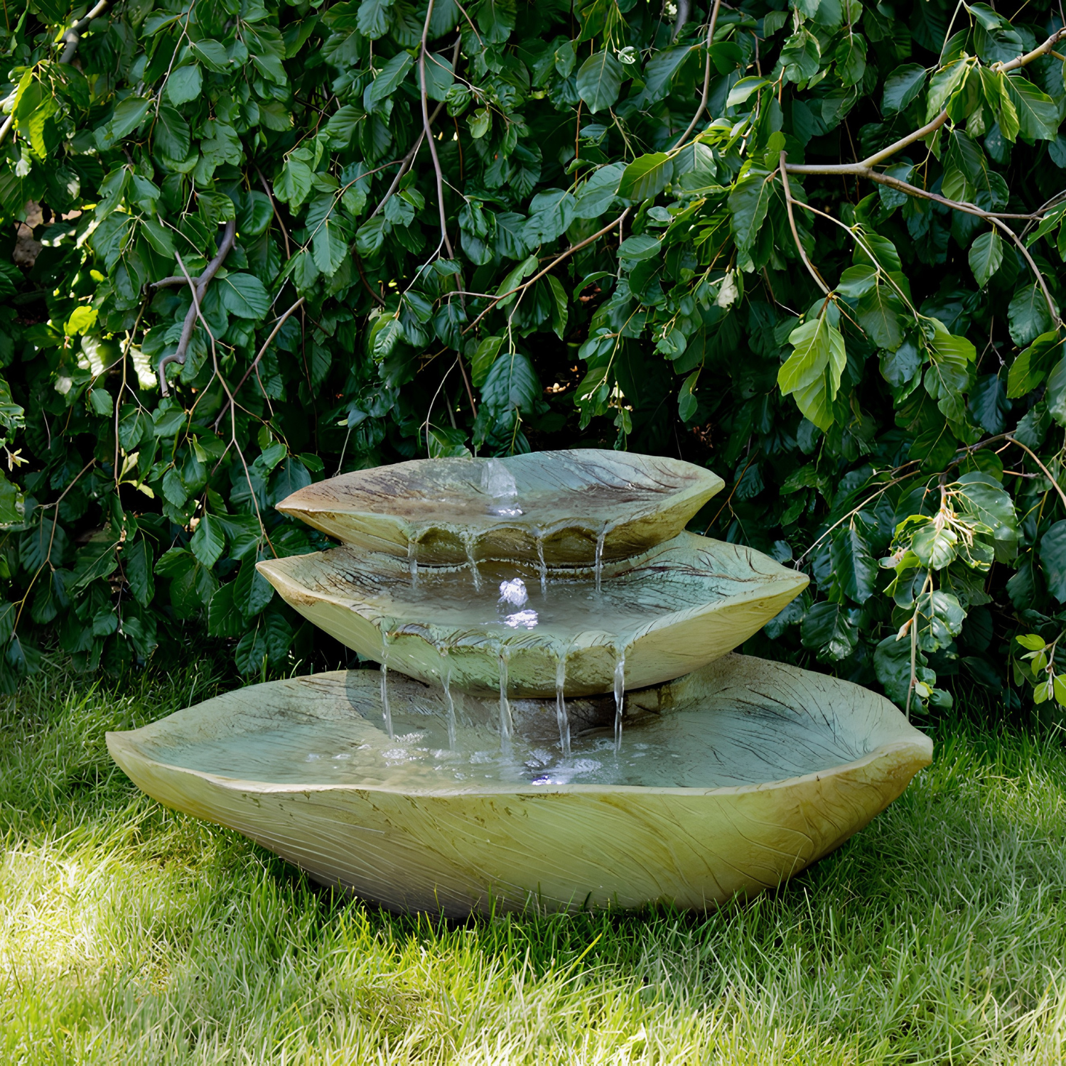 A three-tiered stone fountain with cascading water, set outdoors on green grass in front of a dense leafy hedge.