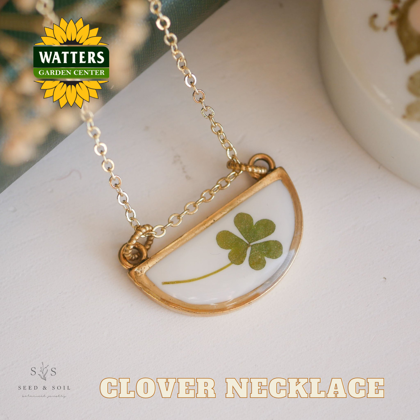 Gold necklace with a clover pendant on a white surface, featuring Watters Garden Center logo.