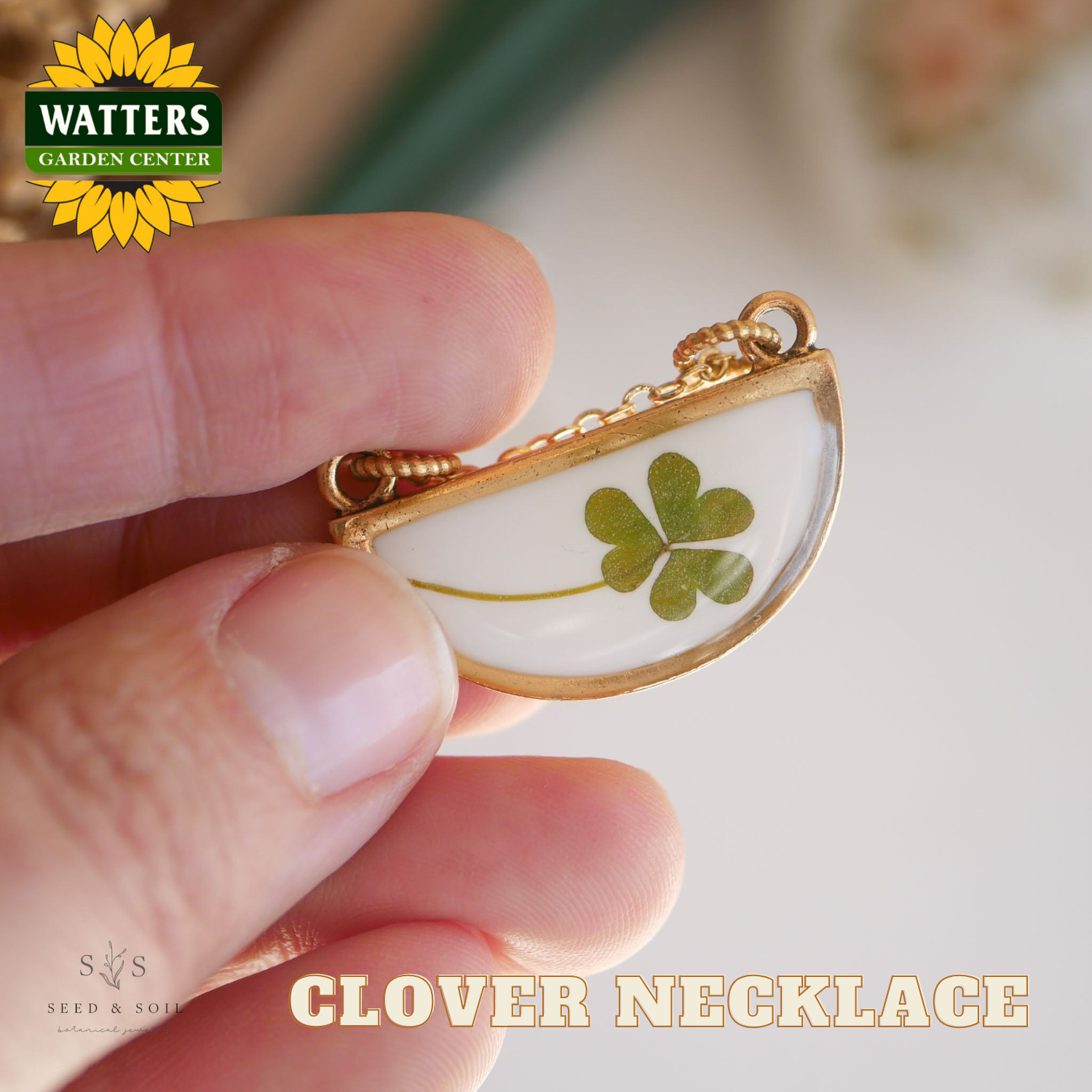 Gold necklace with a clover pendant held by a hand, branded with Watters Garden Center.