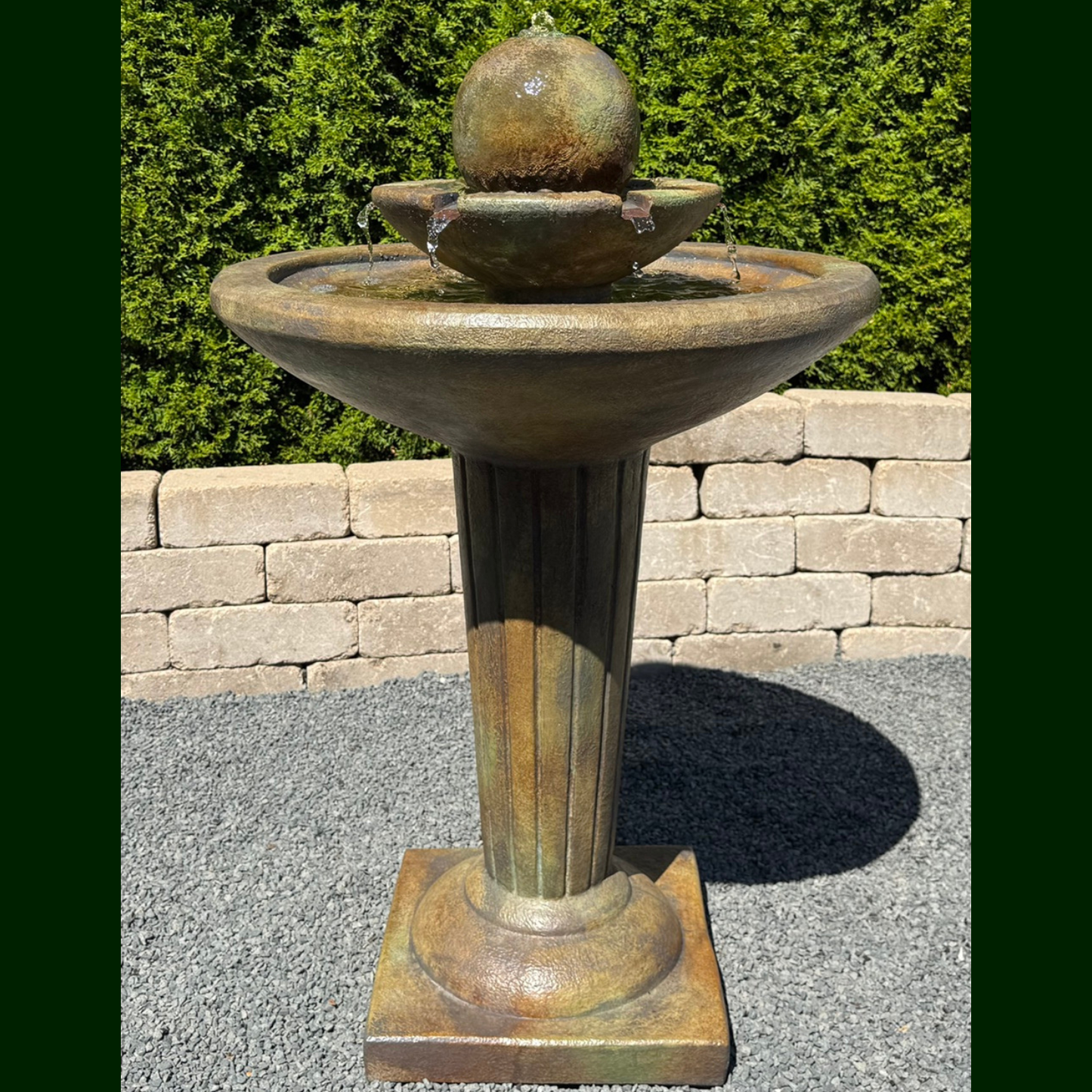An outdoor pedestal fountain with a round stone-like top, shallow upper basin, and fluted column on a square base, set on gravel with a stone wall and green hedge behind it.