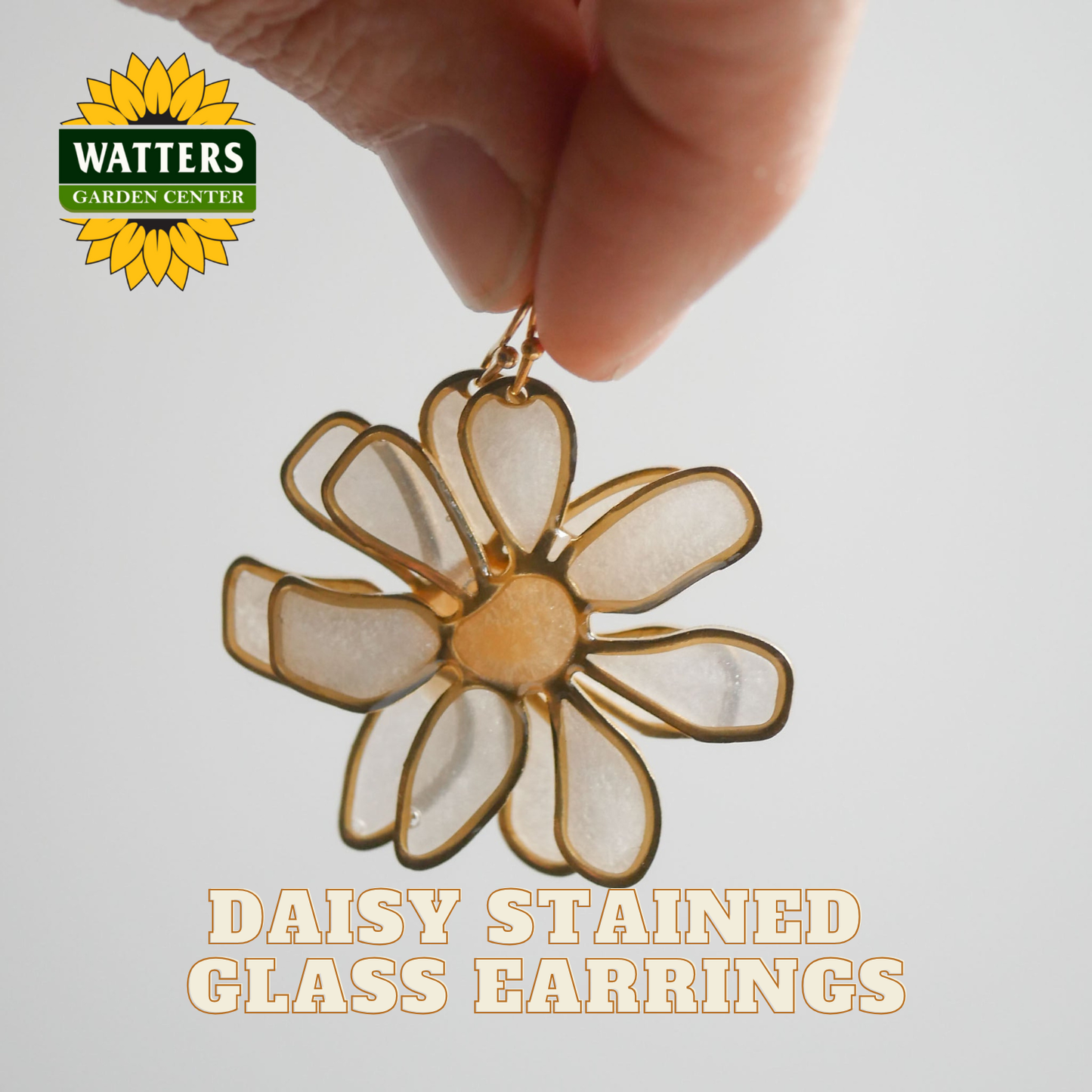 Daisy stained glass earring held by a hand with Watters Garden Center logo.