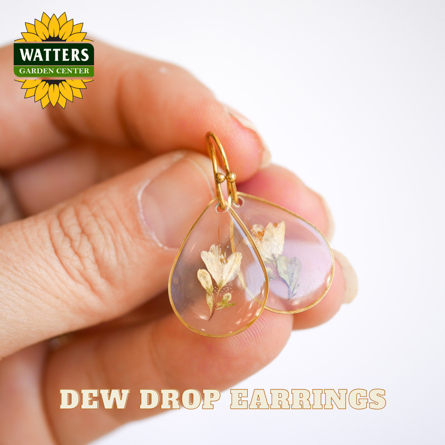 Gold dew drop earrings with floral design held between fingers, Watters Garden Center logo visible.
