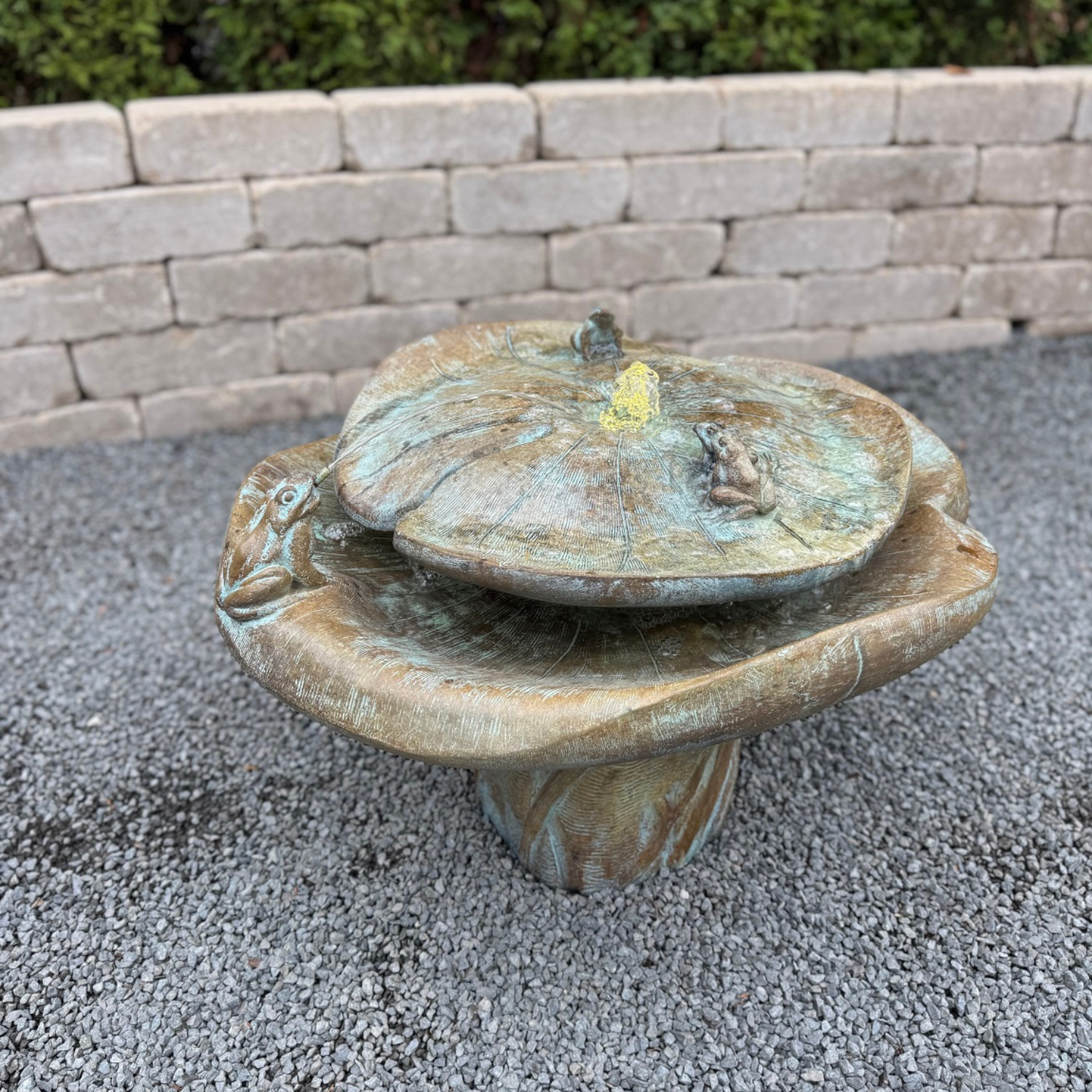 A stone garden fountain shaped like a lotus flower with a frog perched on the rim, set on gravel in front of a light stone wall.