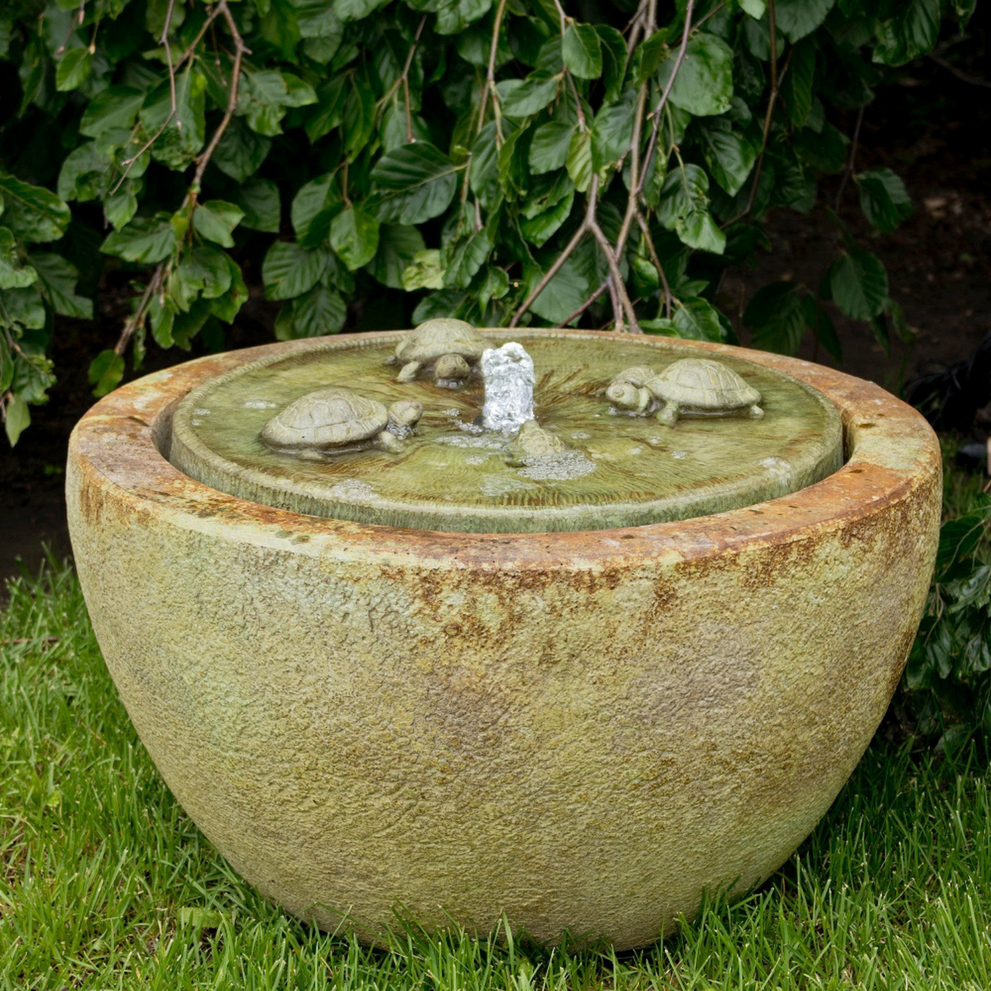 A round stone garden fountain with a textured beige and green surface, featuring a lid with sculpted turtle figures and a central water spout, set on grass in front of leafy green plants.