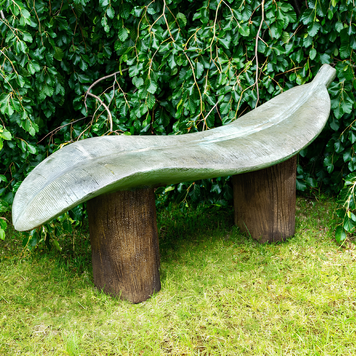 A leaf-shaped outdoor bench made of weathered gray-green stone or concrete, supported by two dark brown cylindrical posts, set on grass in front of a dense green hedge.