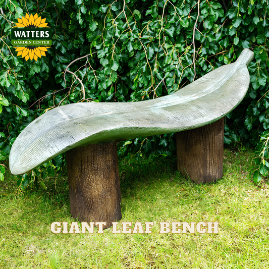 A leaf-shaped outdoor bench made of weathered gray stone or concrete, mounted on two dark brown cylindrical posts, set in a garden with green grass and a leafy hedge background. Text overlays read 'GIANT LEAF BENCH' and a 'WATTERS GARDENS CENTER' logo is visible in the top left.