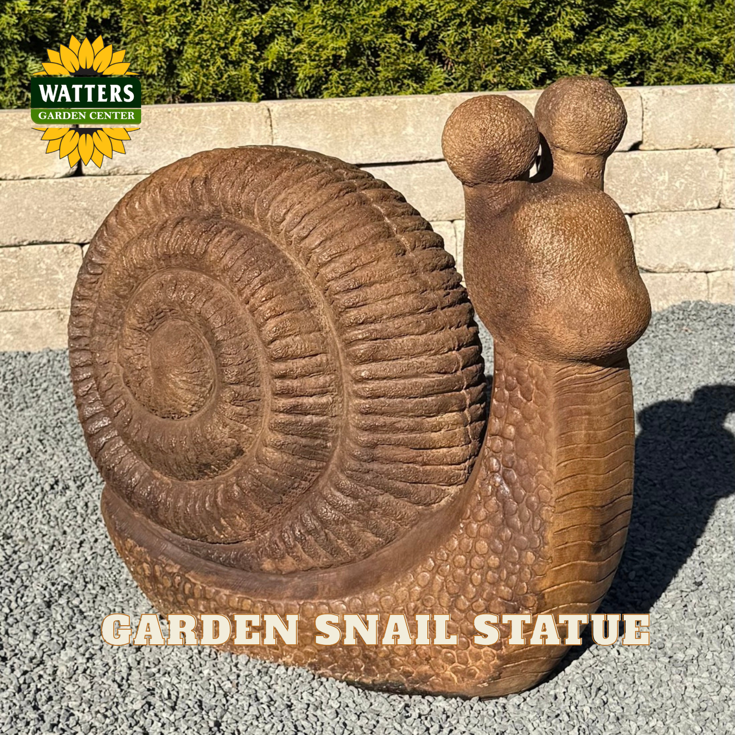 A large realistic garden snail statue with a textured brown shell and body, standing outdoors on gravel in front of a stone wall and greenery. A Watters Garden Center logo appears in the top left, and the text 'GARDEN SNAIL STATUE' is overlaid at the bottom.