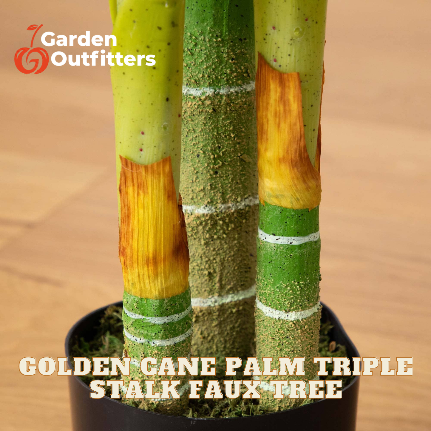 Close-up of a potted artificial palm trunk decoration labeled 'Garden Outfitters' and 'GOLDEN CANE PALM TRIPLE STALK FAUX-TREE,' showing three faux stalks in green, yellow, and orange tones in a black planter on a light wood surface.