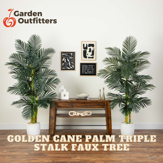 Two tall artificial golden cane palm trees in white planters flanking a wooden console table against a light wall, with framed wall art above and Garden Outfitters branding and product text visible.