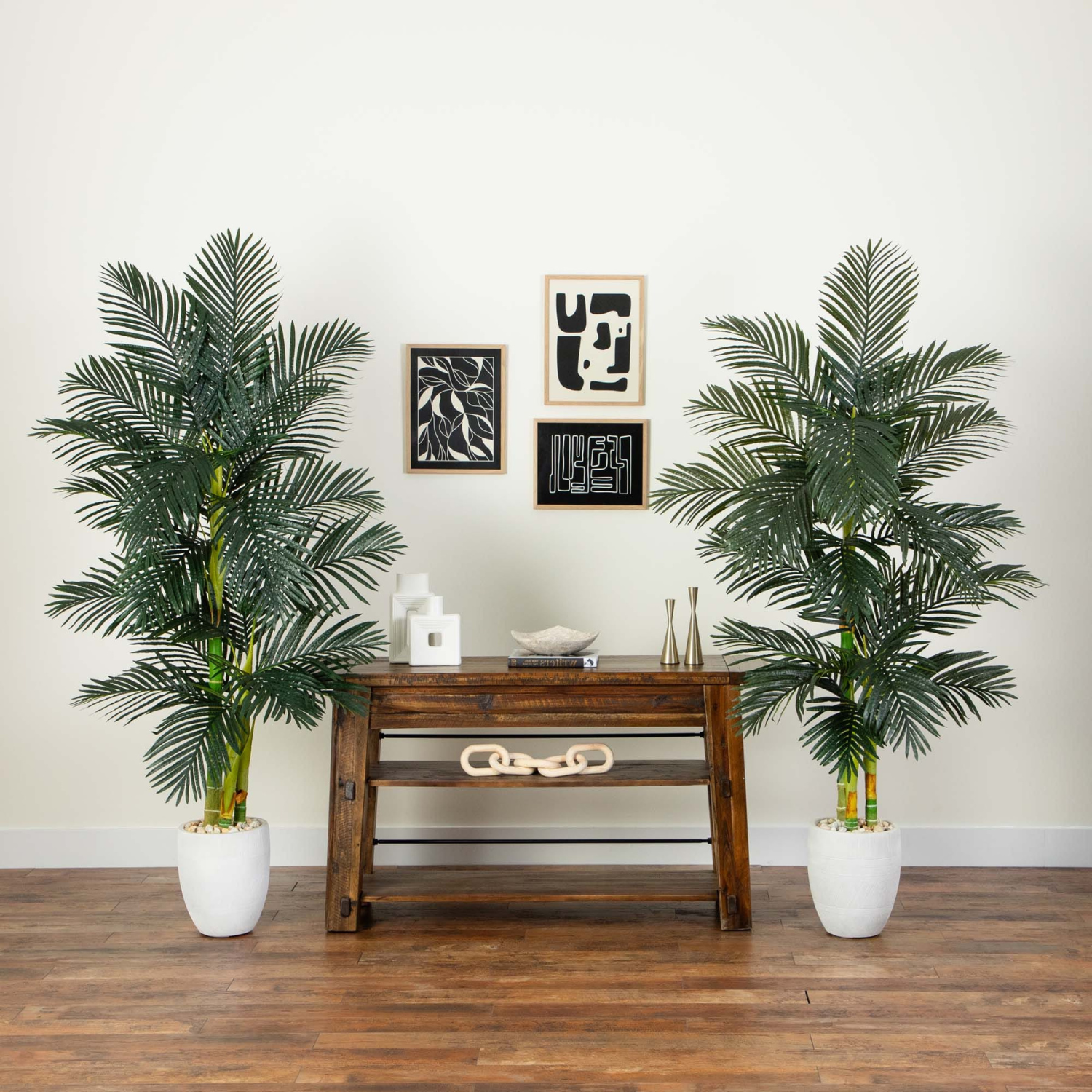 Two tall artificial palm trees in white planters flanking a dark wood console table against a white wall with framed abstract art.