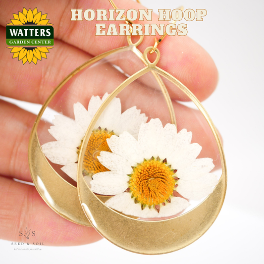 Gold hoop earrings with white flowers held by a hand, branded with Watters Garden Center.