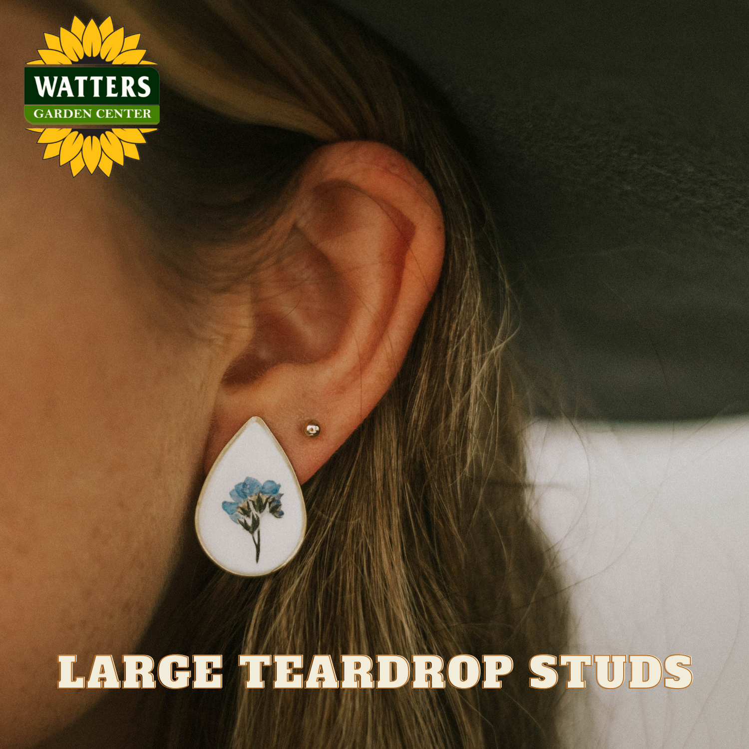 Teardrop-shaped earrings with floral design worn by a person, with Watters Garden Center logo.