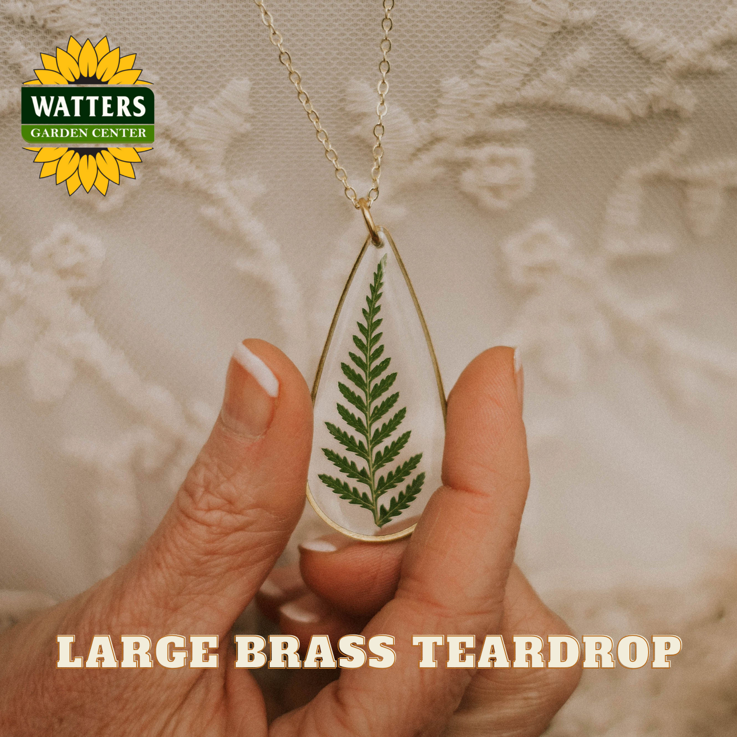 Large brass teardrop necklace with a leaf design held by a hand, Watters Garden Center logo visible.