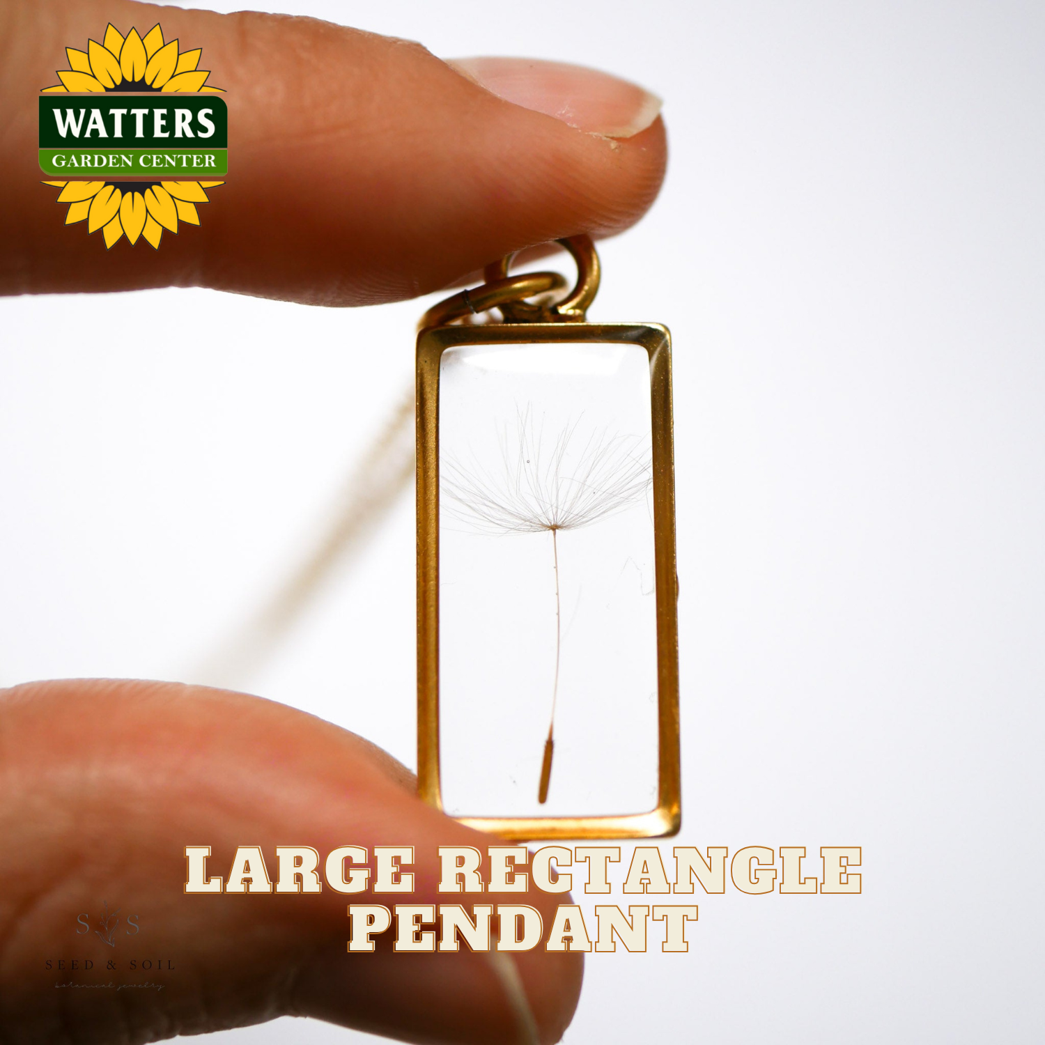 Gold pendant with dandelion seed head held between fingers, Watters Garden Center logo visible