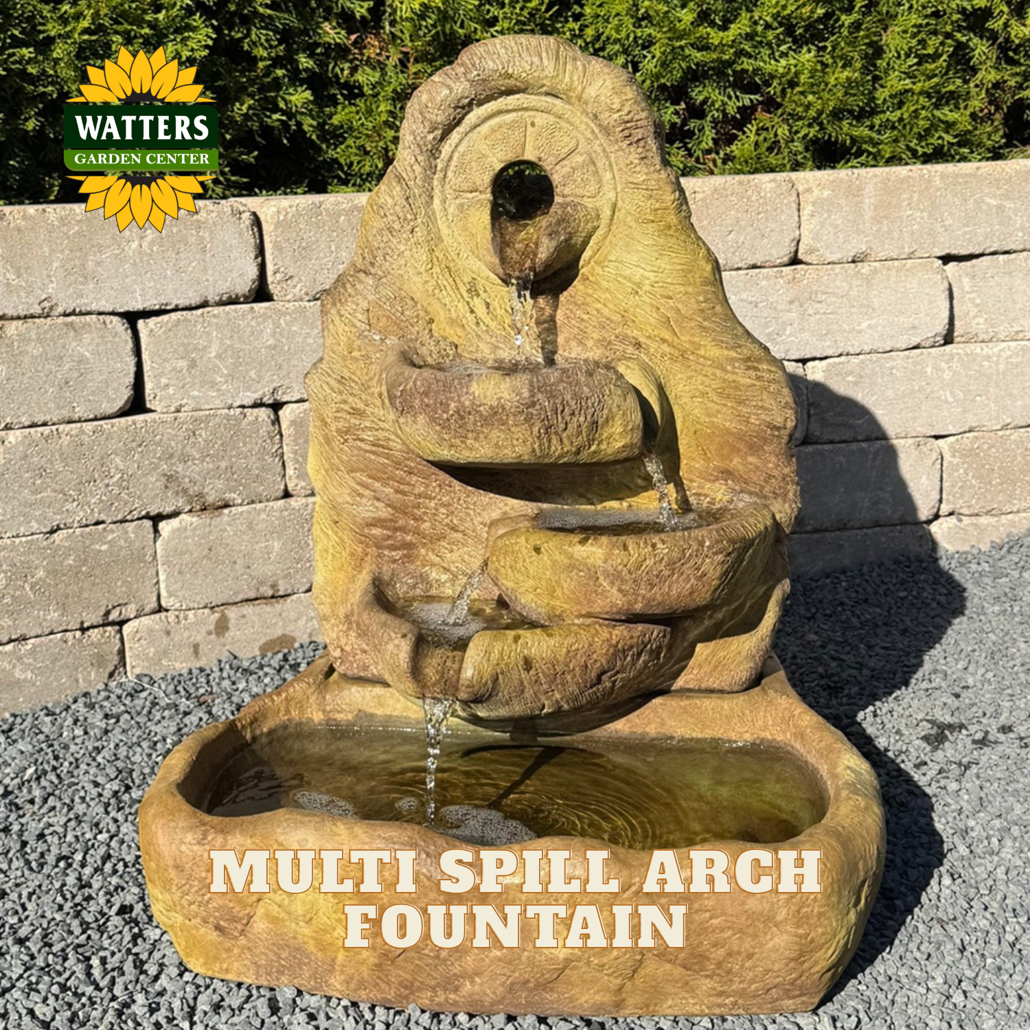 A stone-look multi spill arch fountain with water flowing from an upper opening into curved tiers and pooling in a basin below. The fountain is photographed outdoors on gravel in front of a stone wall and greenery, with the text 'MULTI SPILL ARCH FOUNTAIN' overlaid at the bottom and a Watters Gardens Center logo at the upper left.