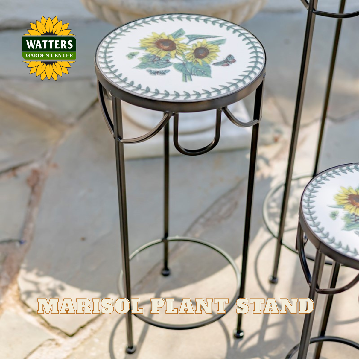 A Watters Gardens Center Marisol plant stand with a round white ceramic tile top featuring a sunflower and green leaf border, supported by a black metal frame, shown outdoors on a stone patio.