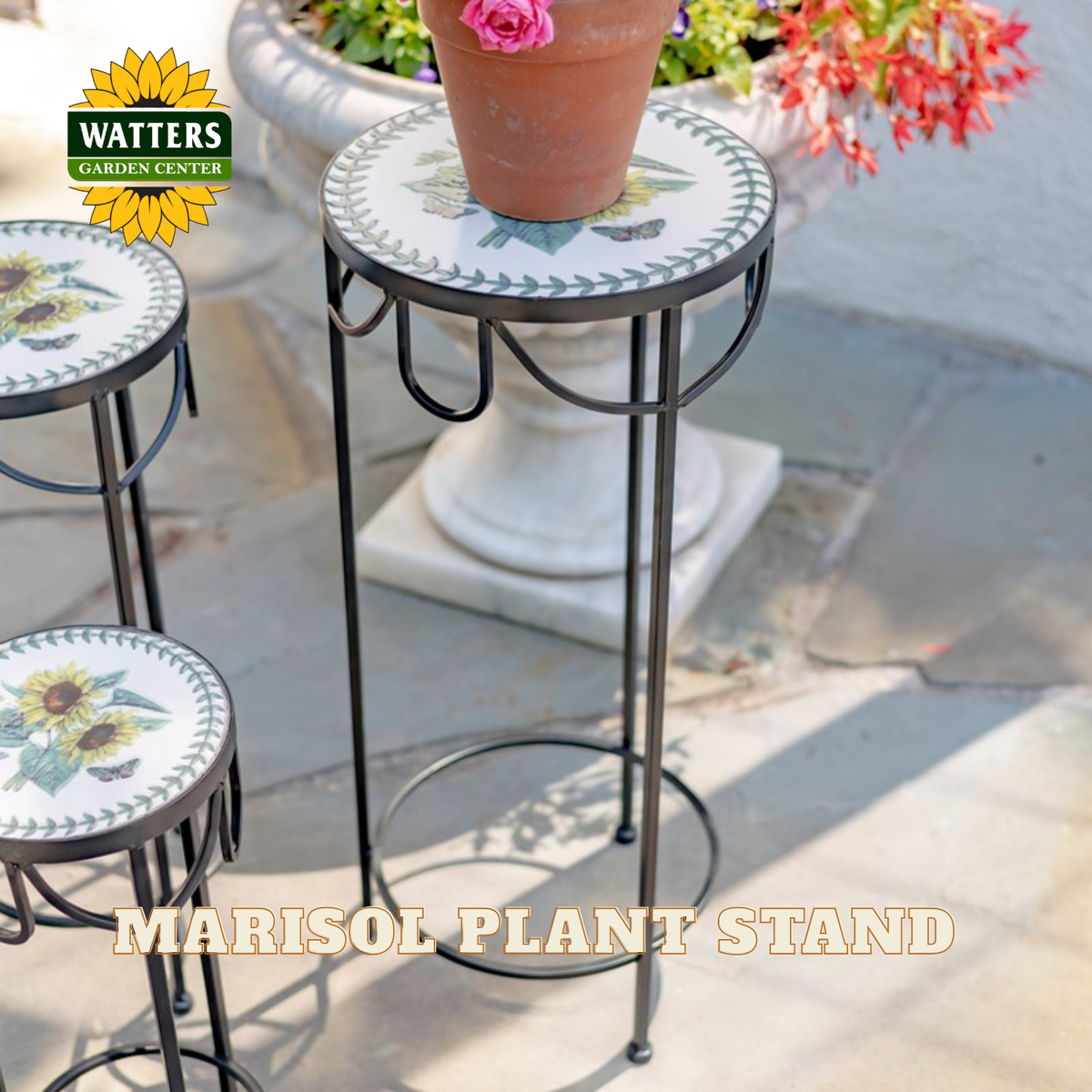 A black metal plant stand with a round mosaic tile top featuring sunflowers and green leaves, shown outdoors on a stone patio. The stand is labeled 'MARISOL PLANT STAND' and a 'WATTERS GARDEN CENTER' logo is visible in the top left corner.