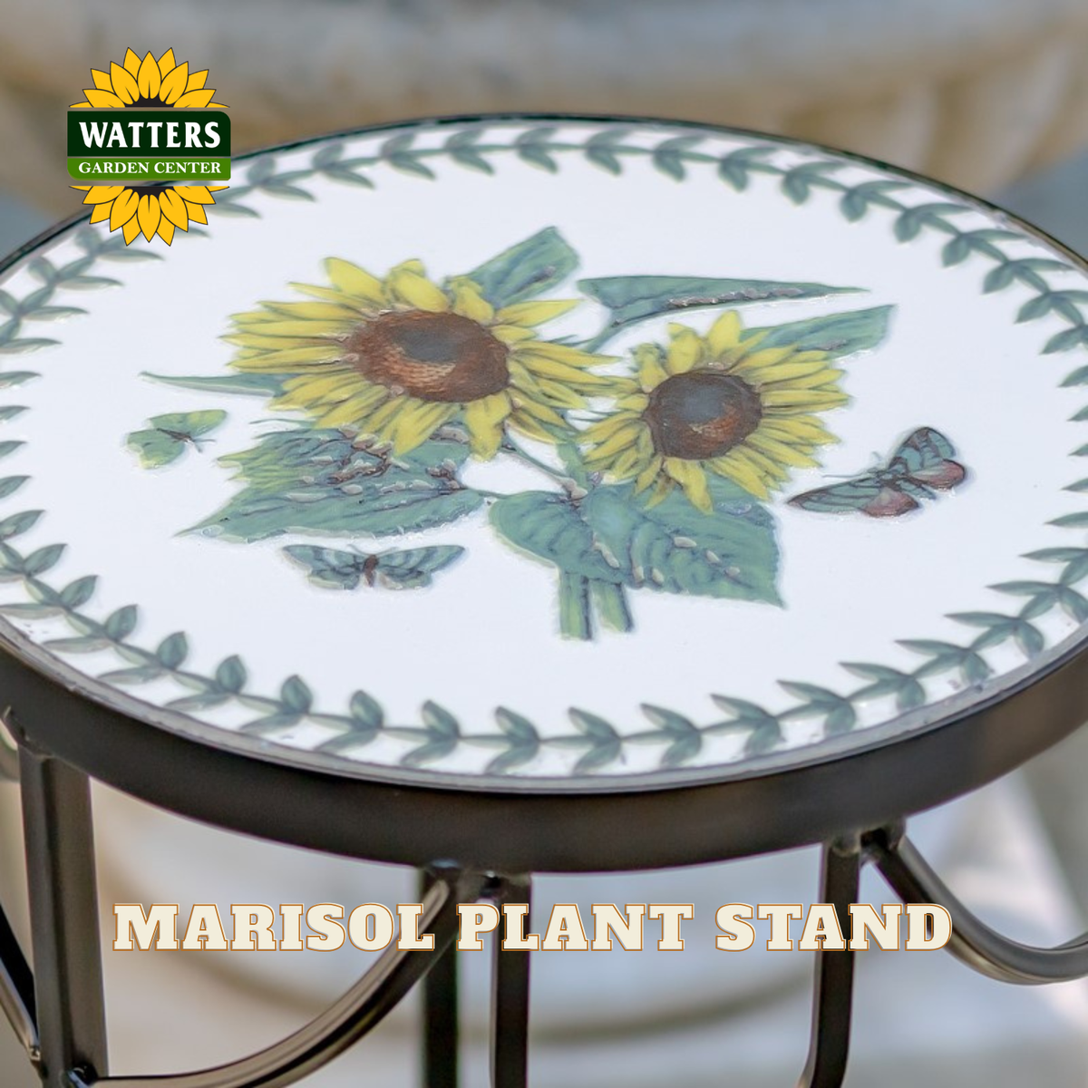 A Watters Gardens Center Marisol plant stand with a white circular top featuring two yellow sunflowers, green leaves, and butterflies, surrounded by a green leaf border. The stand has a dark metal frame and is shown with a logo overlay in the top left and the text 'MARISOL PLANT STAND' at the bottom.