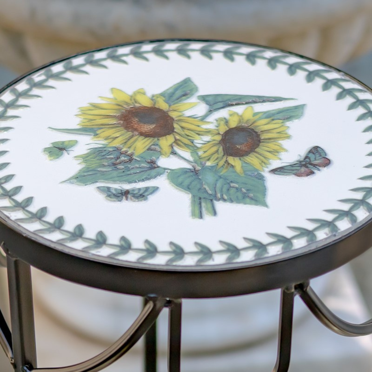 Round outdoor side table with a white ceramic top featuring a yellow sunflower and green leaf border design, supported by a dark metal frame.