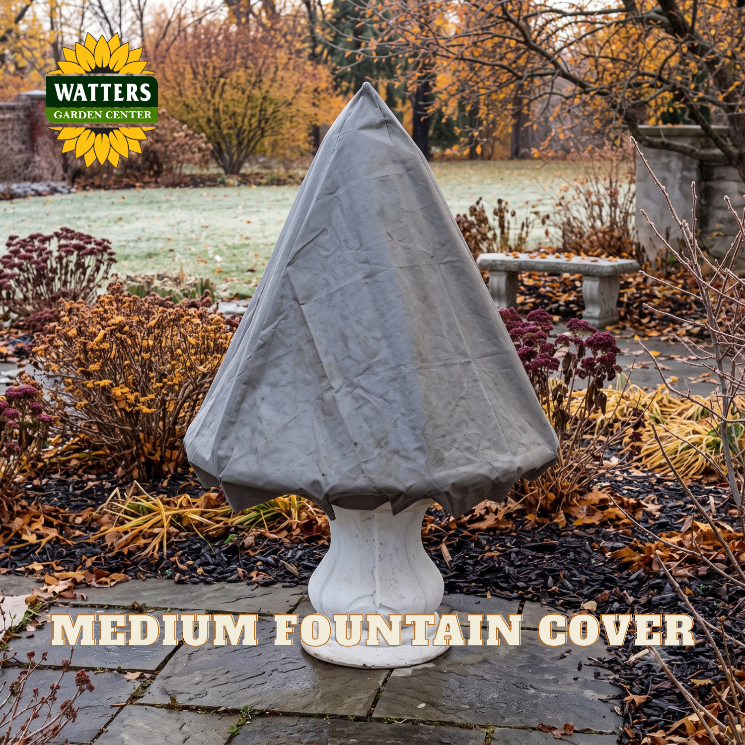 A gray fabric fountain cover fitted over a white stone fountain base, photographed outdoors in a garden setting with fallen leaves, a bench, and trees. The Watters Gardens Center logo appears in the upper left, and the text 'MEDIUM FOUNTAIN COVER' is overlaid at the bottom.