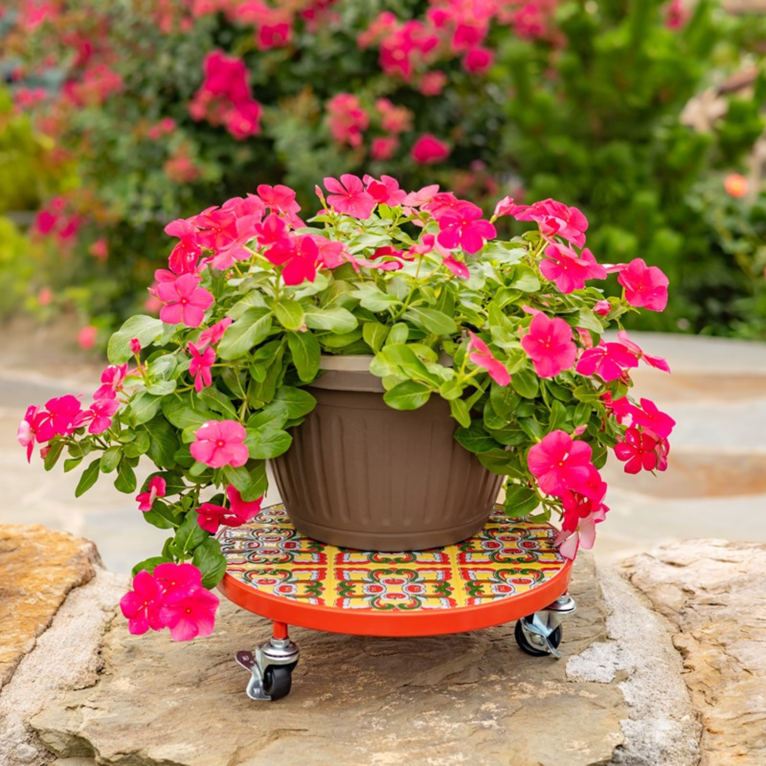 A potted flowering plant with bright pink blossoms and green foliage sits on a decorative orange rolling planter with a multicolor mosaic tile top, placed outdoors on a stone surface with a blurred garden background.