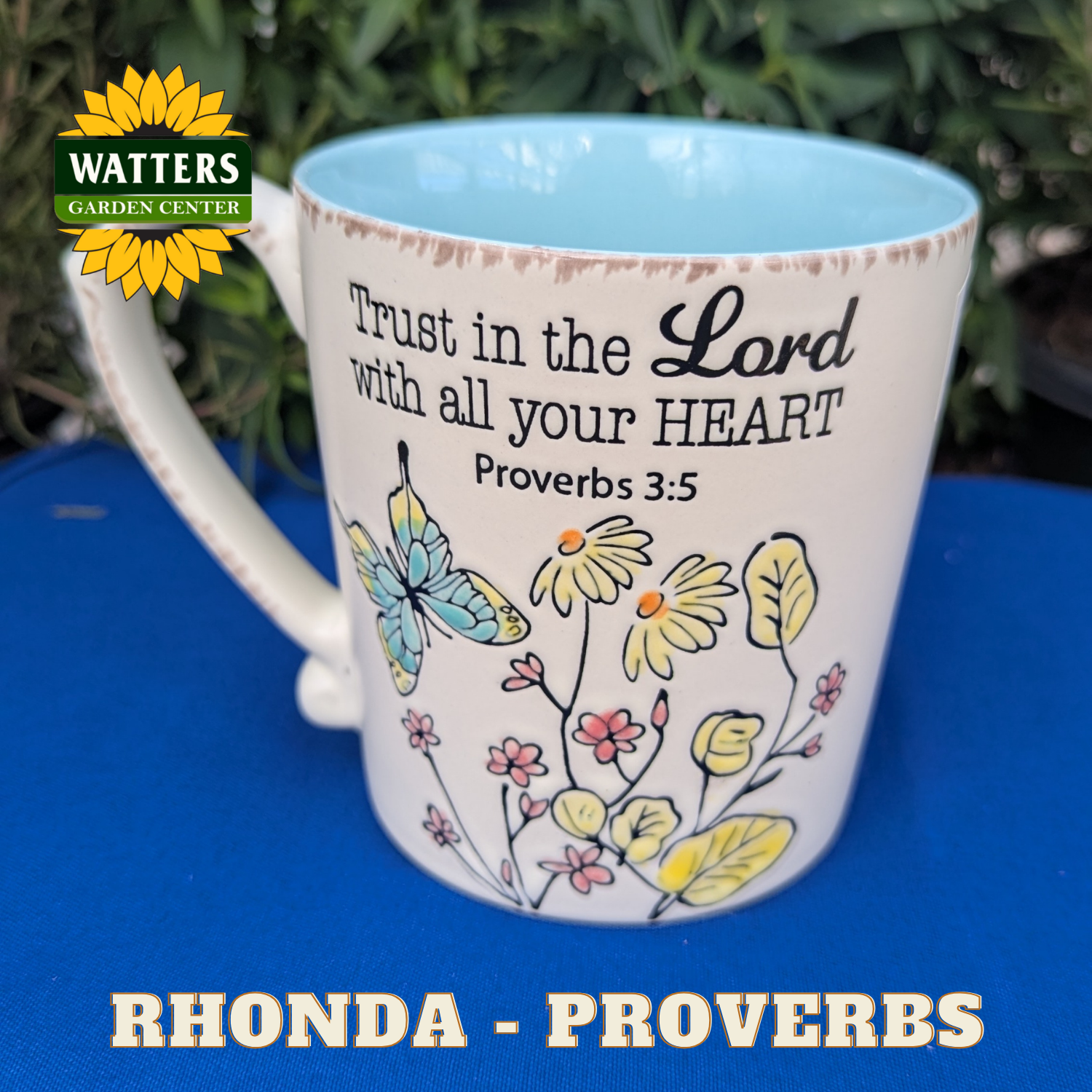 Mug with floral design and motivational quote on a blue surface, featuring Watters Garden Center logo.