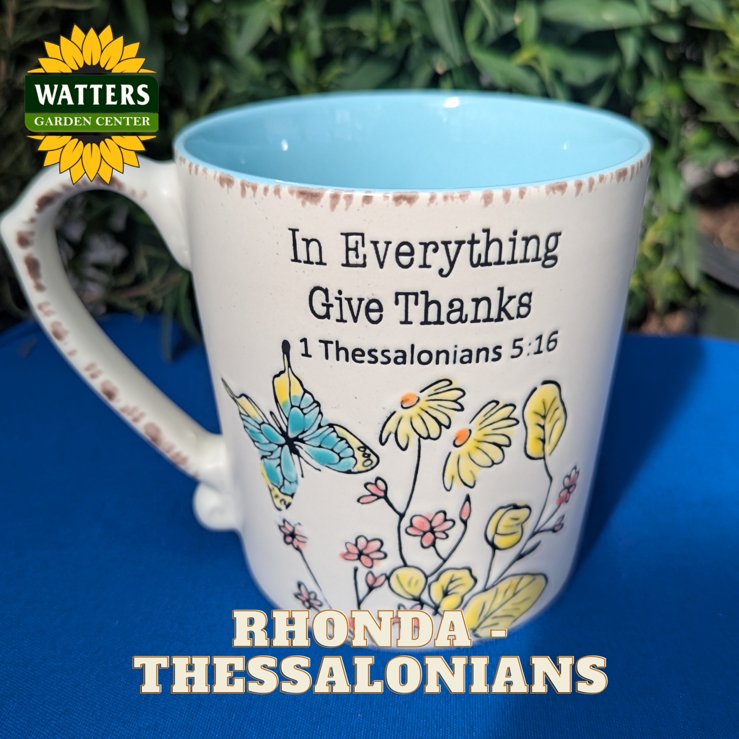 Mug with floral design and text 'In Everything Give Thanks 1 Thessalonians 5:16' on a blue surface with Watters Garden Center logo.