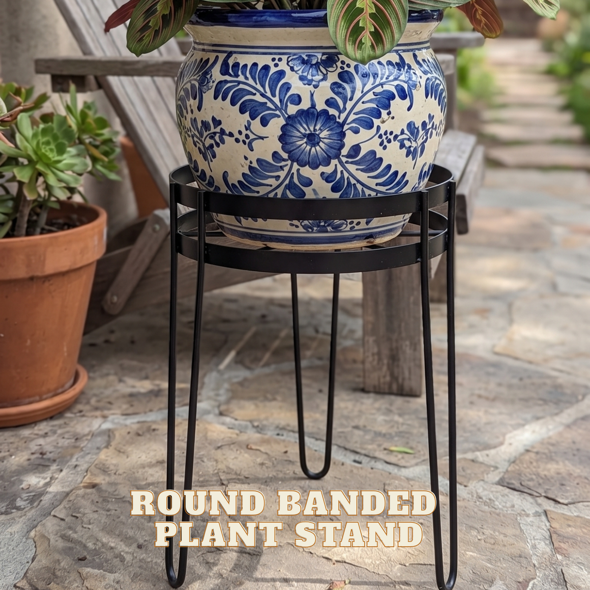 A round banded plant stand with a black metal hairpin frame, holding a blue and white floral ceramic planter. The stand is photographed outdoors on a stone patio next to other potted plants, with the text 'ROUND BANDED PLANT STAND' overlaid at the bottom.