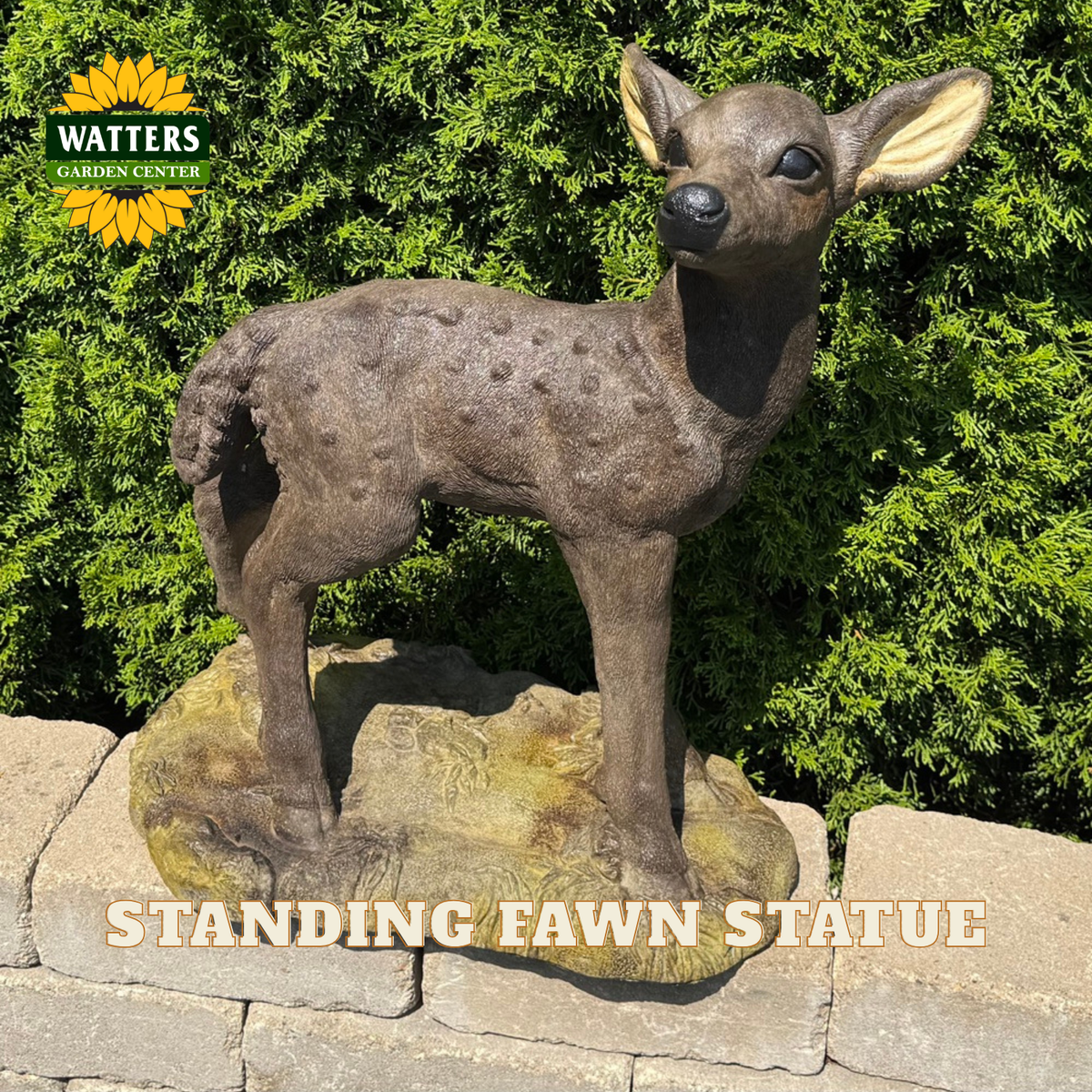 A realistic standing fawn statue with a spotted brown coat and light tan ears, mounted on a textured rock base. The statue is photographed outdoors in front of dense green shrubs, with a stone wall below.