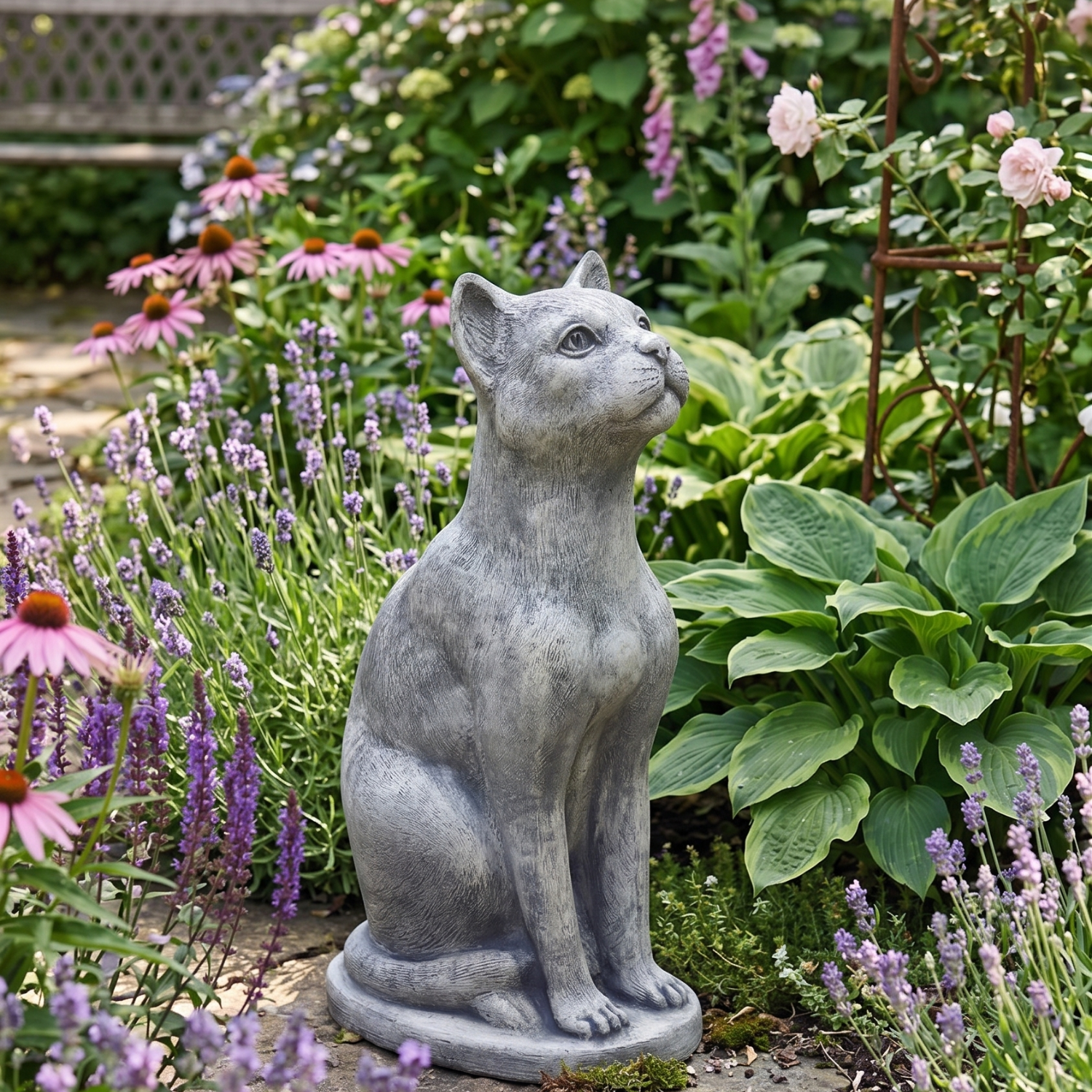 A gray stone-look cat garden statue sitting upright in a lush garden bed surrounded by pink flowers and green foliage.