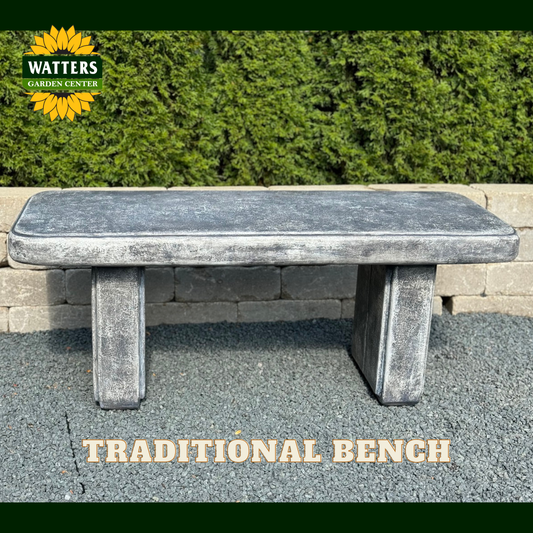 A rectangular stone bench with a weathered gray finish, set outdoors on a paved surface in front of a green hedge. The Watters Garden Center logo is visible in the top-left corner, and the text 'TRADITIONAL BENCH' is displayed at the bottom.