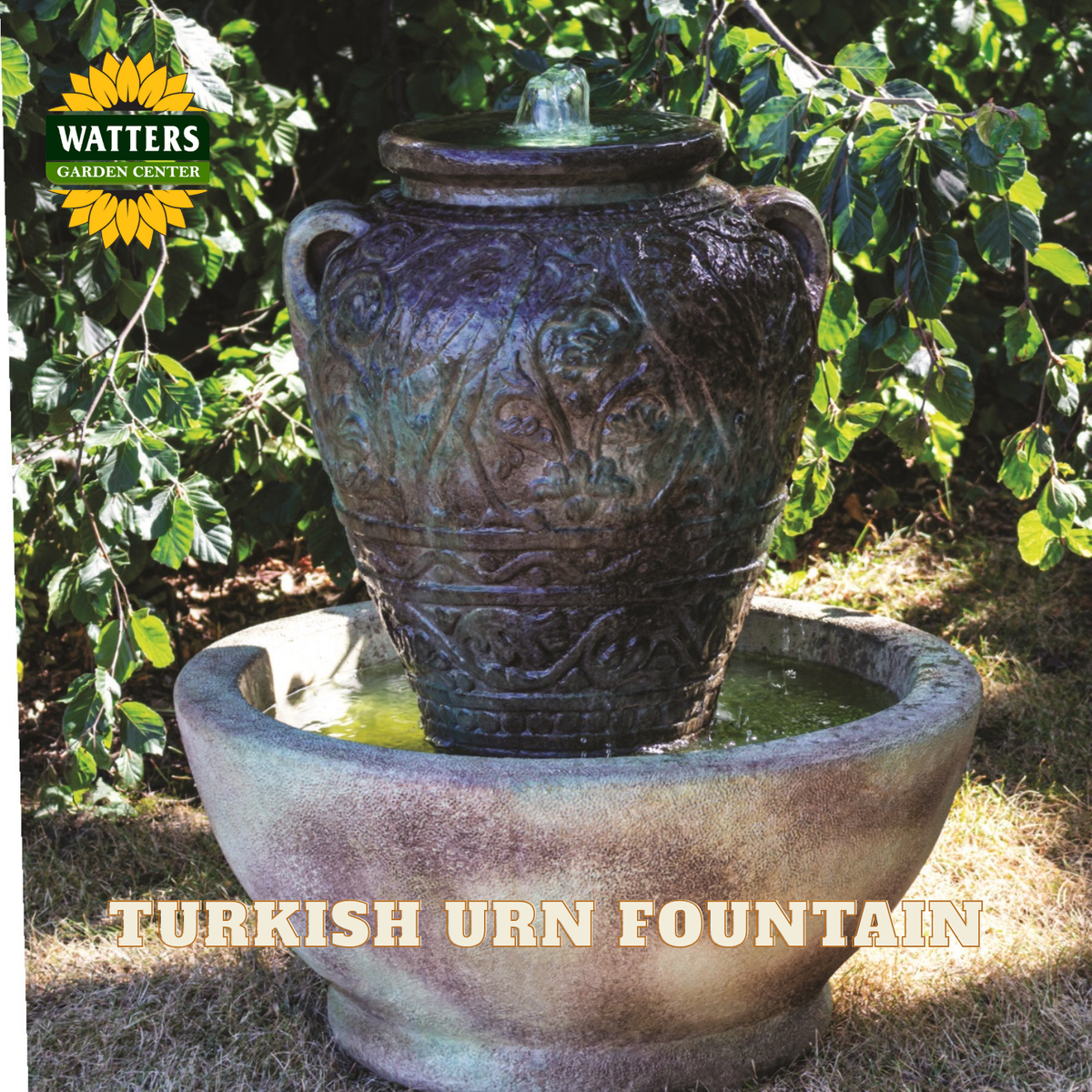 A Watters Garden Center Turkish Urn Fountain shown outdoors in a garden setting. The fountain features a dark, textured ceramic urn with carved details, with water flowing from the top into a matching stone basin below.