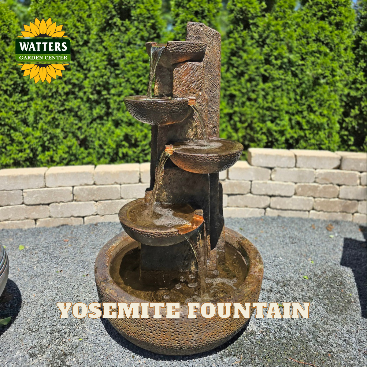 A rustic three-tier stone fountain with water flowing from top to bottom, set outdoors on a gravel surface with a stone wall and green hedges behind it. The Watters Garden Center logo appears in the top-left corner, and the text 'Yosemite Fountain' is overlaid at the bottom.