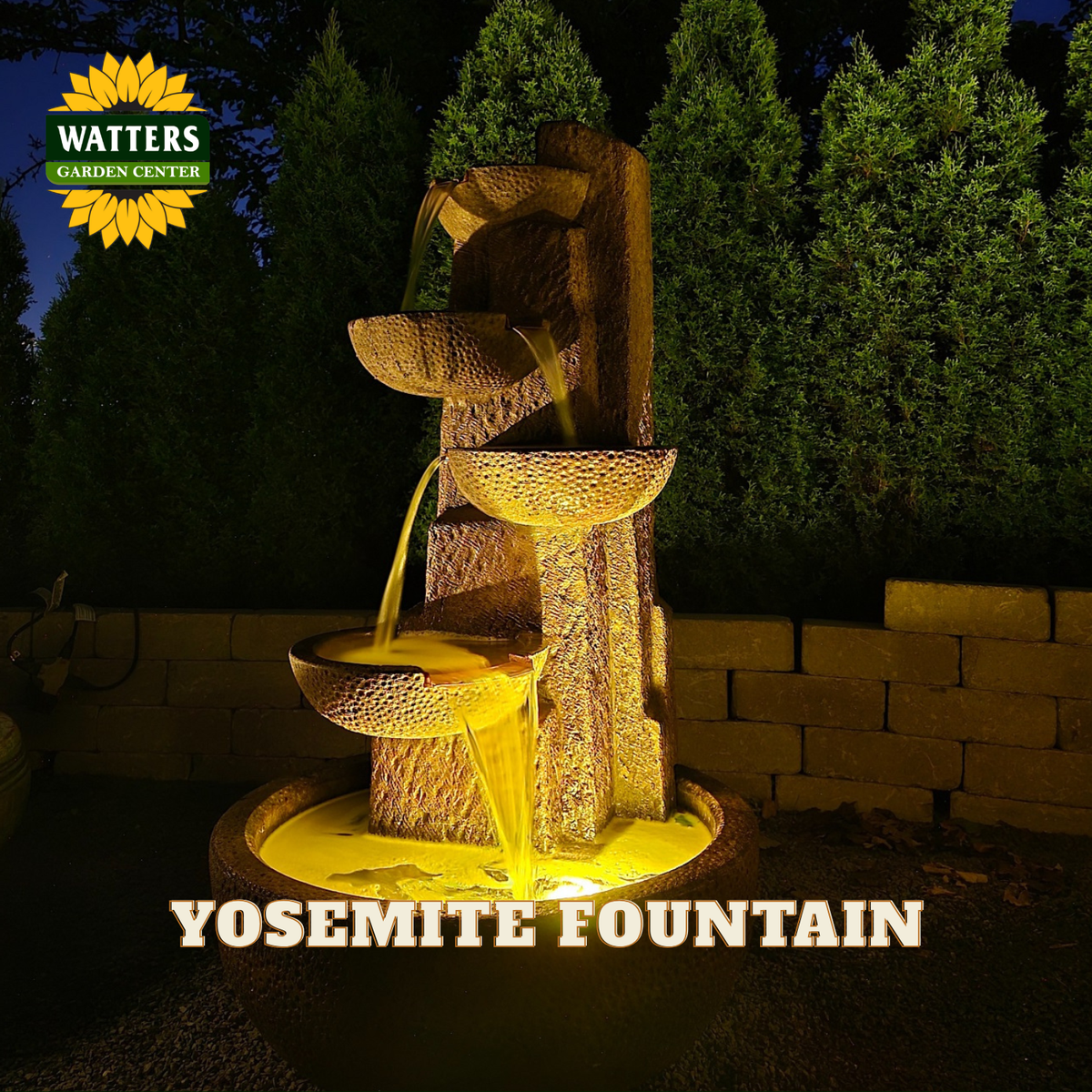 A tiered outdoor waterfall fountain with a textured stone-like finish, illuminated by warm yellow lighting. The fountain has three levels with flowing water and a round basin at the bottom.